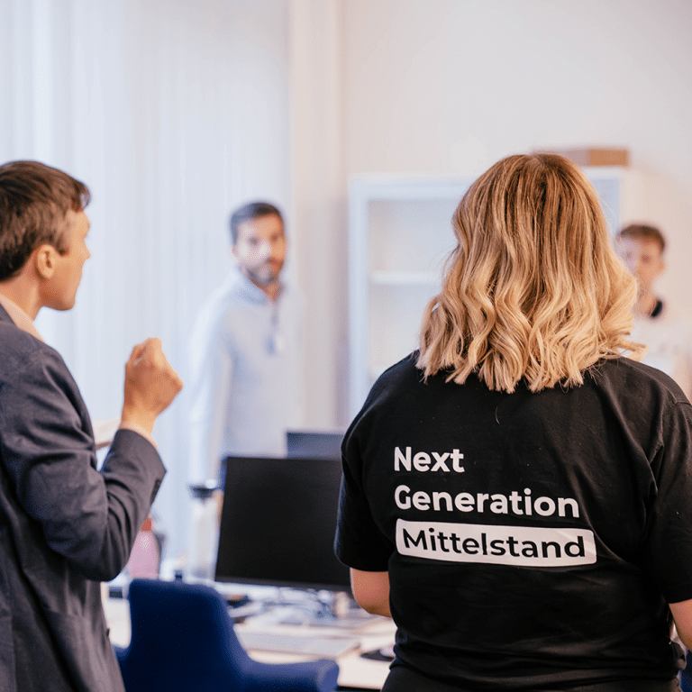 Zu sehen sind mehrere Personen in einer Besprechung - prominet zu sehen ist ein T-Shirt-Rücken mit der Aufschrift "Next Generation Mittelstand".